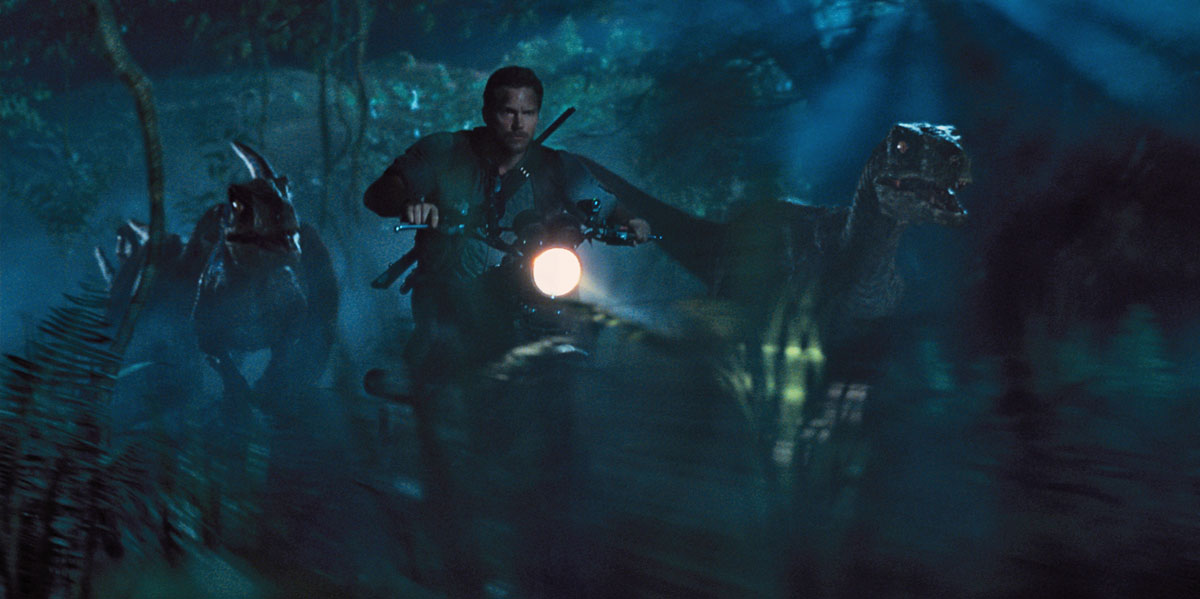 Chris Pratt on a motorbike riding away from dinosaurs, in Jurassic World.