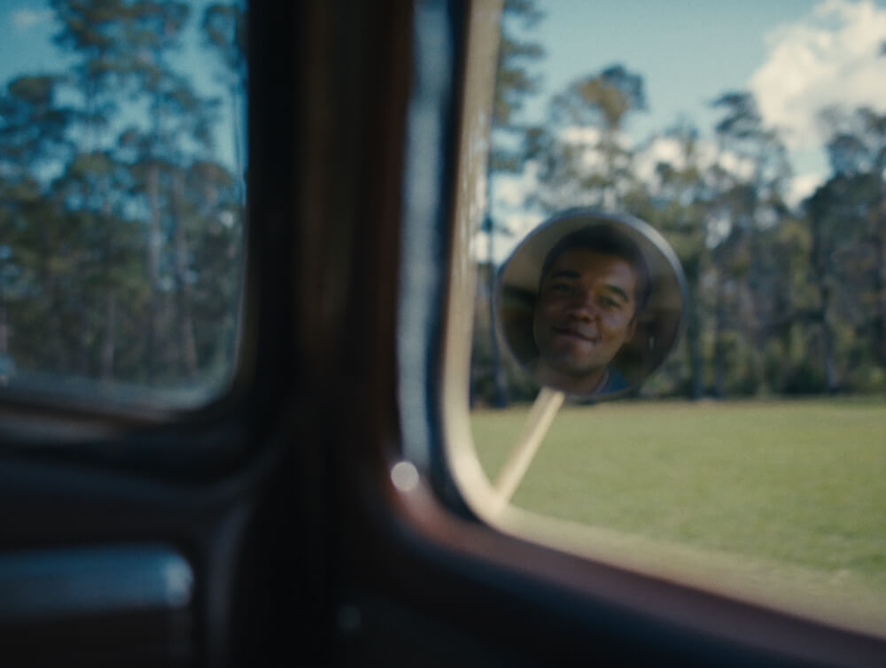 A reflection of a young man in a car's side mirror shows a smirk and contemplative look into the distance, with trees and a field visible in the background