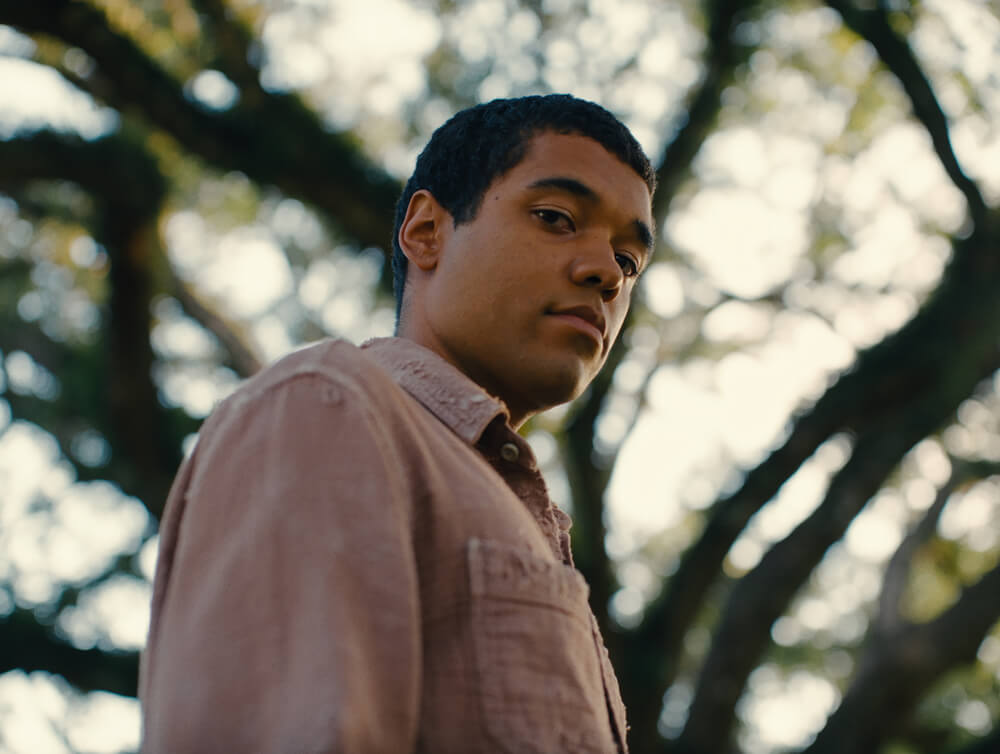 Actor Brandon Wilson (as Turner) gazes contemplatively to the side, shot from a low angle with sunlight filtering through blurred trees in the background.