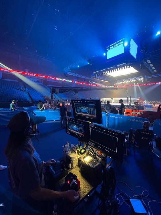 Behind-the-scenes view of cinematographer Rina Yang, BSC monitoring a boxing arena, while blue stage lighting illuminates the space and empty stadium seating.