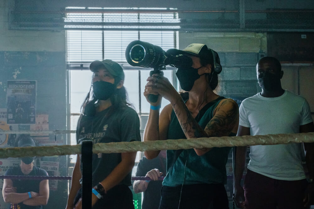 Cinematographer Rina Yang, BSC (left) and director Rachel Morrison, ASC stand behind a boxing ring rope, with Morrison holding a directors viewfinder with telephoto lens attached in a window lit boxing gym