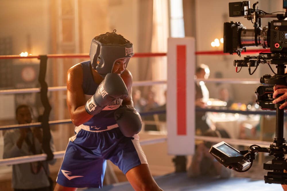 Ryan Destiny as gold-medalist boxer Claressa Shields in blue shorts and protective headgear takes a fighting stance in a boxing ring, while a professional camera and crew film the scene