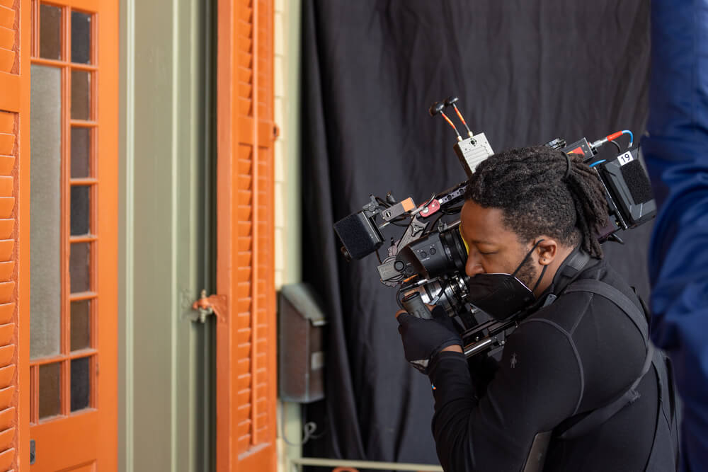 Cinematographer Jomo Fray operates a handheld camera rig as he captures a shot through an orange-painted door with multiple window panes